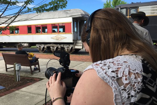 Woman with brown hair stands behind a camera at a train station