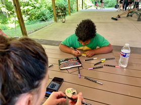 An adult and a young person sit at a wooden picnic table crafting ornaments for the U.S. Capitol Christmas Tree. Markers are spread out on the table.