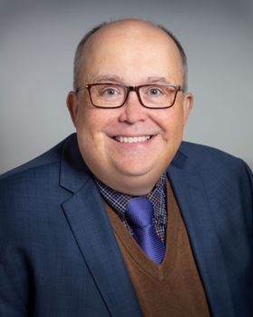 Headshot of Potomac State College president Cris Gilmer. He is pictured in front of a light gray background and is wearing a navy blue jacket with a brown, v-neck sweater, plaid dress shirt and purple tie. He is balding and wearing square-framed glasses. 