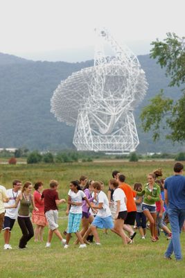 A large group of school aged children run and play in a grassy area with the giant Green Bank telescope in the background. 