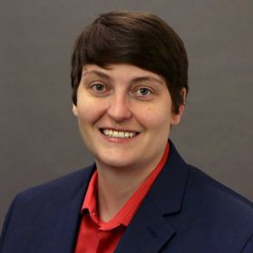 This is a portrait of Jill Gibson. She is sitting in front of a gray background wearing a red shirt and blue jacket.