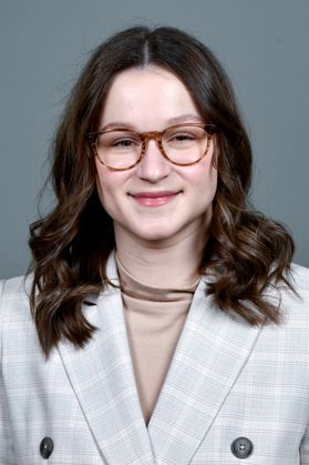 Headshot of WVU Bucklew Scholar Rose Puzzini. She is pictured in front of a gray background and is wearing a gray plaid jacket over a taupe dress shirt. She has long, auburn hair and is wearing round framed glasses. 