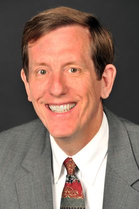 Headshot of Mark Koepke. He is pictured against a gray background and is wearing a gray suit with a white shirt and red, gray, and blue necktie. He has short light brown hair and is smiling in the photo. 