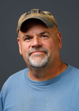 A man with a camp hat and a blue shirt smiling