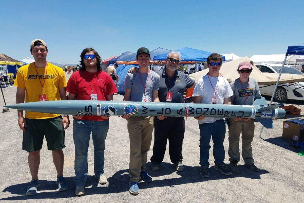 six males holding a large blue tube in front three mountain peaks