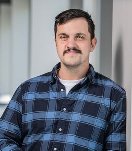 Headshot of economist Zachary Porreca. He is pictured leaning against a beige wall wearing a blue plaid shirt. He has short black hair and a trimmed black mustache. 
