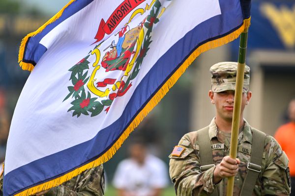 A photograph of a soldier dressed in fatigues holding a flagpole with the state of West Virginia flag blowing in the wind. 