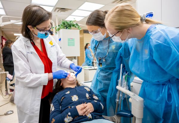 A dentist wearing a white coat wearing a white mask and blue latex gloves works on a young patient who is wearing a blue sweatshirt. Two dental students in blue gowns observe on the right side of the photo.