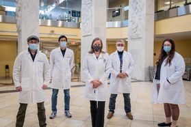 people in lab coats, masks stand in large room 