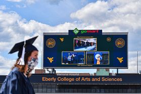 woman in cap, gown and masks walks in front of a scoreboard on football field