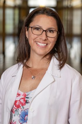 Headshot of WVU researcher Tatiana Trejos. She is wearing a floral top under her white lab coat. She has shoulder length brown hair and is wearing glasses. 