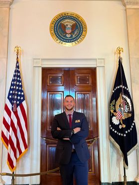 WVU student veteran Ben DeRoos is show here outside of the Oval Office during his attendance at the Student Veterans of America Leadership Institute in Washington DC. He is wearing a dark suit and has his arms crossed in front of him. 