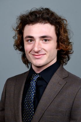Headshot of WVU Bucklew Scholar Michael Groh. He is pictured against a gray background wearing a brown jacket over a black dress shirt with a dark patterned tie. He has shoulder length curly brown hair. 