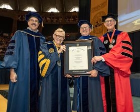 WVU Faculty Senate Chair Scott Wayne, WVU President Gordon Gee and Davis College Dean Darrell Donahue present Joel Newman with an honorary degree in a brown frame. All are in academic regalia.