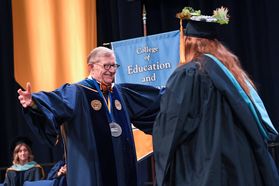 man in glasses greets student in cap and gown with outstretched arms