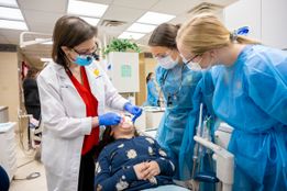 Photo of a child in a dental exam chair. A female dentist in a white coat is working with instruments in the child's mouth while two female dental students observe. 