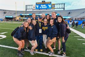 A group of eleven students huddle for a picture on the Milan Puskar Stadium field. 