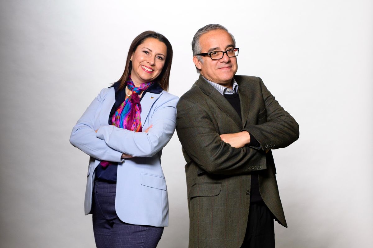 Two people stand back to back in front of a white backdrop. One is wearing a light purple jacket. One is wearing a dark jacket.