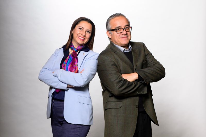 Two people stand back to back in front of a white backdrop. One is wearing a light purple jacket. One is wearing a light jacket.