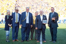 Group of 5 men in suits and one woman in pants and a sweater posing with awards on a football field.
