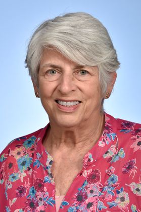 Headshot of Dr. Judith Feinberg. She is pictured against a blue background and is wearing a pink, floral top. She has very short gray hair and is smiling in the photo. 