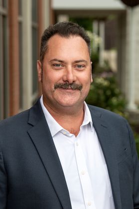 Headshot of Andy Williamson, director of outdoor community development for WVU’s OEDC. Williamson is pictured outside wearing a dark jacket and white dress shirt. He has dark hair and a mustache. 