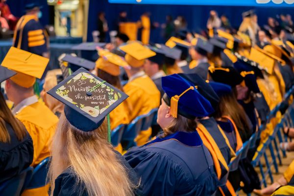 People in gold and blue caps and gowns sit in multiple rows; In the foreground, a woman with long blond hair wears a graduation cap that reads, 'Nevertheless, she persisted.'