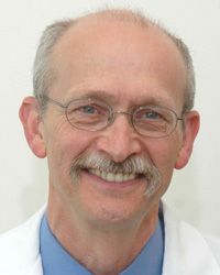 A man looks at the camera while wearing a white doctor's coat. This is a headshot. He has a gray mustache and glasses