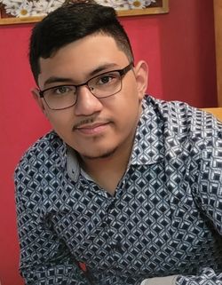 Headshot of WVU student Antonio Perez. He is pictured here seated against a red background. He is wearing a blue patterned dress shirt. He has short black hair and square framed glasses. 