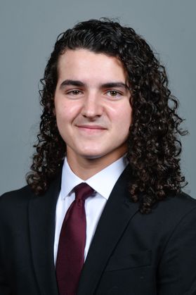 Headshot of WVU Bucklew Scholar Benjamin Blackwell. He is pictured against a gray background wearing a black suit, white dress shirt and maroon tie. He has long, curly dark hair. 