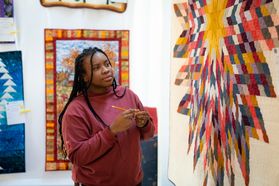 A multi-colored quit in the pattern of a starburst is on a white background while a person in a pink shirt looks at during Mountaineer Week 2021. Other quilts are hanging on the wall behind the person.
