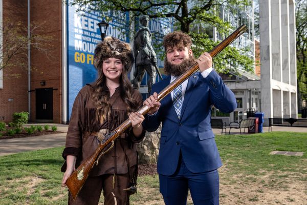 The 68th WVU Mountaineer Mary Roush hands the rifle over to the 69th Mountaineer Mikel Hager during a ceremony on campus. They are pictured outside standing in front of the Mountainlair with Mary in her buckskins and Mikel in a blue suit. 