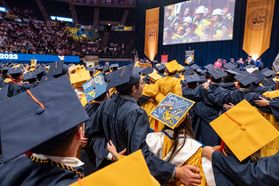Graduates from the Statler College wear gold and blue caps and gowns and stand with their arms around each other while singing "Country Roads."