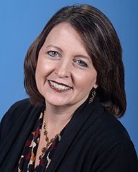 This is a portrait of Amy Snodgrass who has shoulder-length dark hair and is wearing a black jacket with necklace in front of blue backdrop.