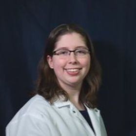 Headshot of WVU researcher Meghan Prusinowski. She is pictured against a dark background wearing a white lab coat. She has shoulder length brown hair and wears glasses. 