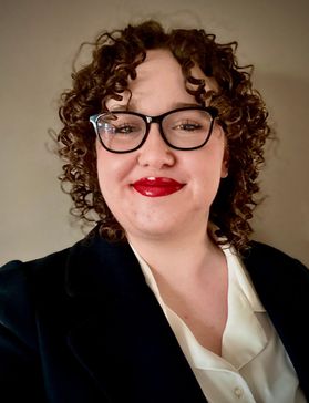 Portrait of Emily Diaz, she wears a dark blazer and glasses. She wears a bright red lipstick and has short curly hair.