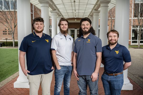 Four Mountaineer mascot candidates pose together at the WVU Erickson Alumni Center. The four males stand centered between white columns and all are wearing WVU-branded attire. 