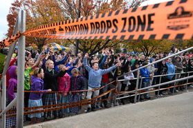 This is a crowd of dozens of students looking up toward the Pumpkin Drop launch point. An orange banner reading 'caution drops zone' stretches across the front of the frame.