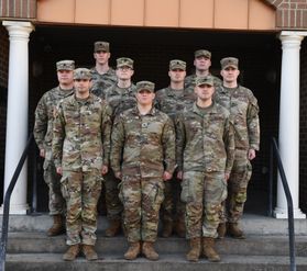 A photograph showing nine WVU Army ROTC cadets standing at attention on cement steps in formation with two white columns on either side of the group. 