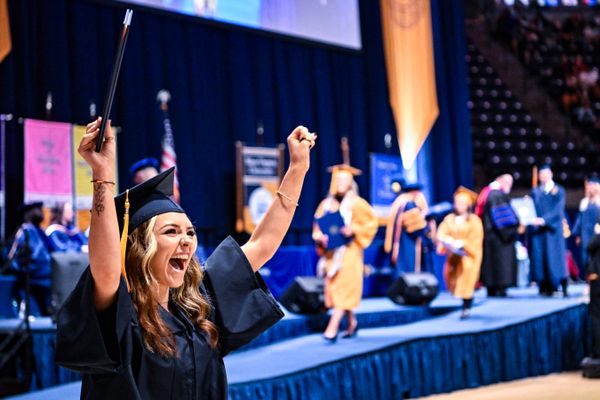 A recent female WVU graduate with long blond hair wearing a navy blue cap and gown holds one fist in the air and her leather bound diploma in the other celebrating her achievement after walking across the Commencement stage.