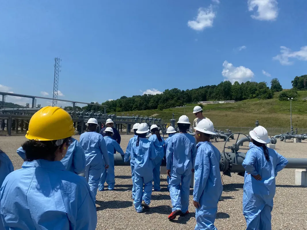 Young children visit a drilling site during a WVU camp. The children are wearing blue safety suits and hard hats while they listen to a male guide. 