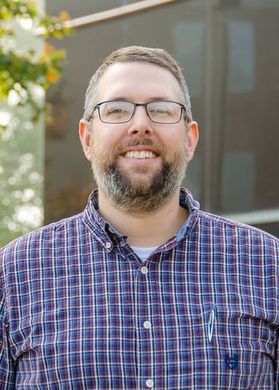 Headshot of Terence Musho, WVU associate professor. Musho is standing outside and is wearing a blue checked shirt. He has a pen in his pocket and is wearing dark-framed glasses. He also has a brown and gray beard. 