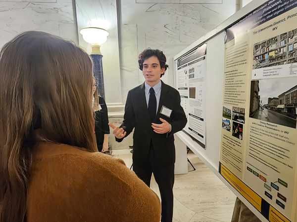A student with short, curly dark hair stands in the rotunda of the State Capitol wearing a dark suit. To his left side is a research poster. A person with long brown hair stands with a back to the camera and listens.