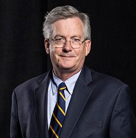 This is a portrait of Fred King. He is wearing a navy blue jacket, gold and blue striped tie and light blue shirt. Fred has gray hair and glasses and is in front of a black background.