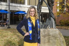 Headshot of WVU student Giana Loretta standing in front of the Mountainlair wearing a gold dress with a blue blazer and a WVU scarf. 