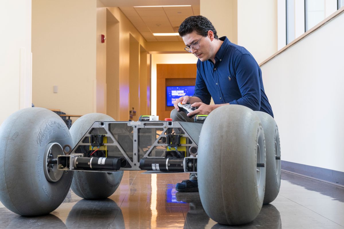 Cagri Kilic, a postdoctoral research fellow, is pictured here wearing a blue shirt, wire-frammed glassed, and with short brown hair. He's using a hand-held remote control to maneuver his rover. 