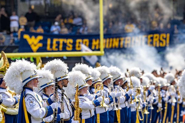 A picture of the WVU Marching Band performing during a football game. There is a blue and gold banner behind them that says 