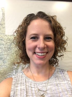 Headshot of WVU emloyee Traci Knabenshue. She is standing in front of a wall map of West Virginia. She has short curly light brown hair and is wearing a beige and white patterned shirt. 