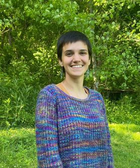 Headshot of WVU Fulbright Scholar Katherine Adase. She is pictured outside with green grass and trees in the background. She is wearing a multi-colored marbled sweater and has very short black hair. 