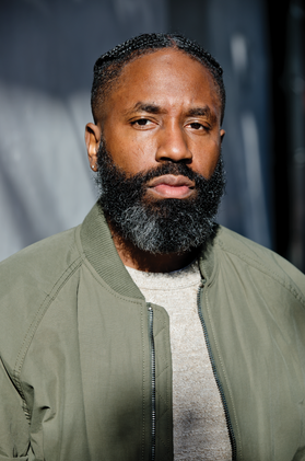 Headshot of Rodney Williams. He is pictured in front of a gray background and is wearing an olive jacket over a gray shirt. He has very short black hair and a long black beard. 